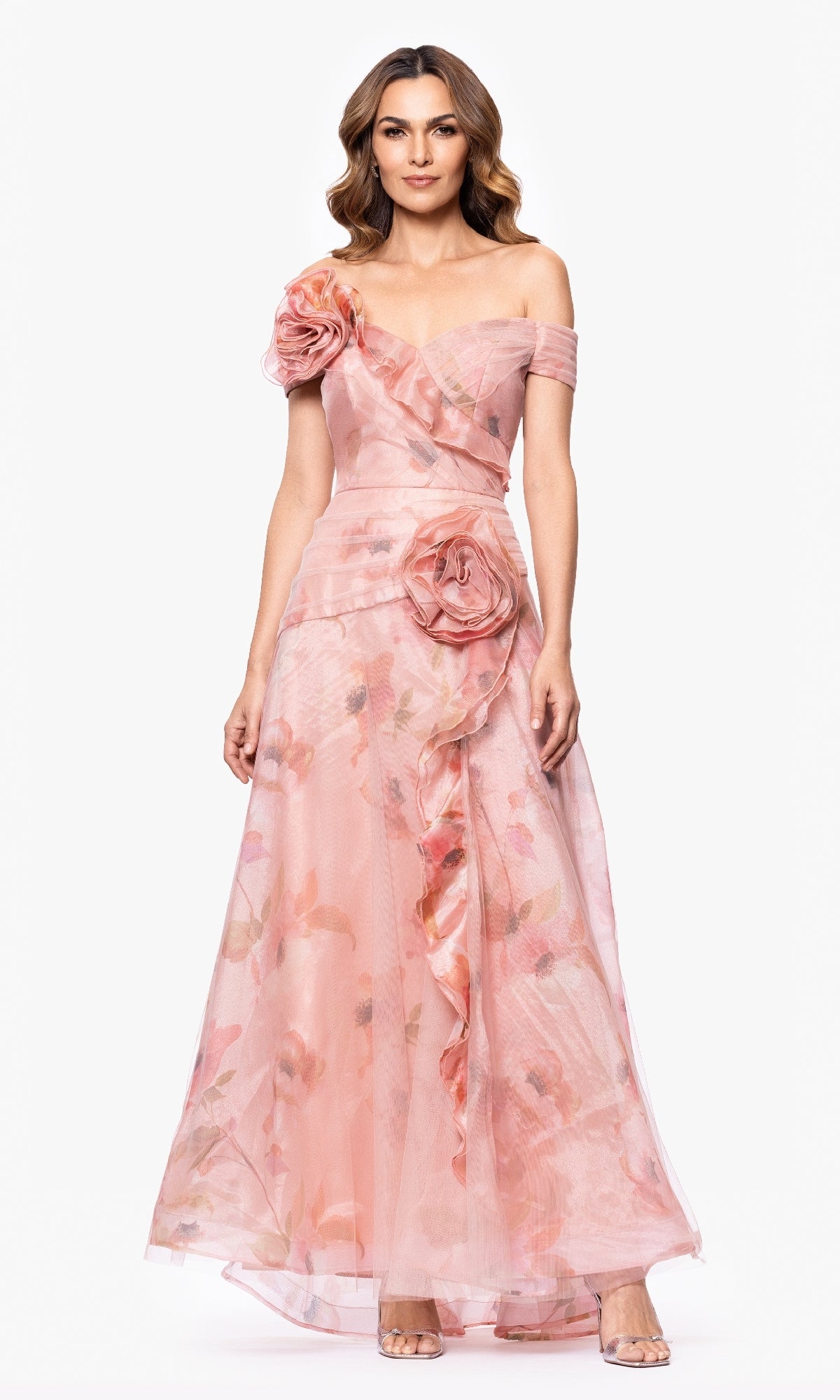 A woman models the Betsy & Adam A27103 long formal dress featuring an off-the-shoulder pink floral print, large fabric flower accents, and an elegant A-line skirt, standing against a plain white background.