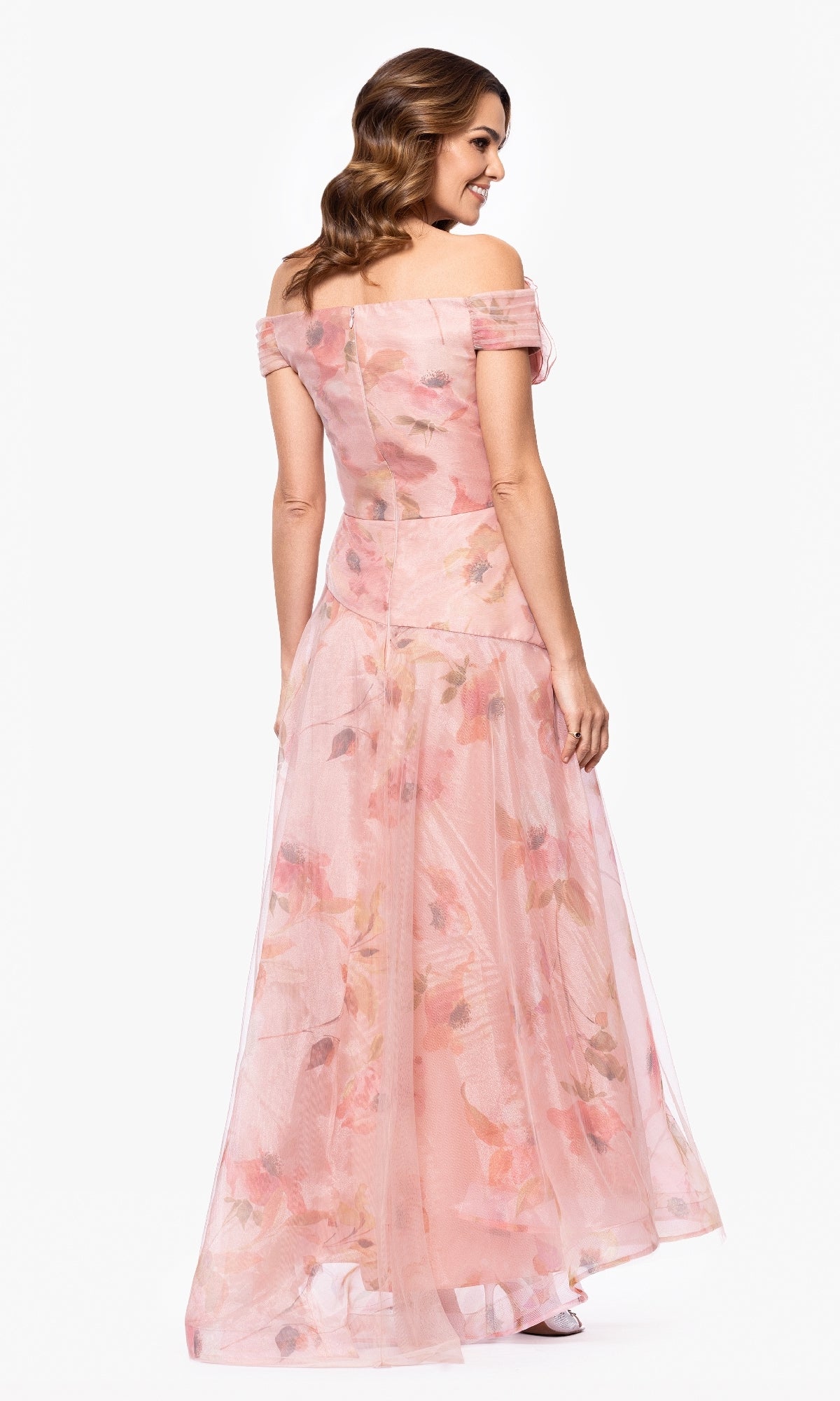 A woman stands facing away in the Betsy & Adam A27103 long formal dress, featuring a floral print, off-the-shoulder neckline, floor-length A-line skirt, and sheer overlay, set against a plain white background.