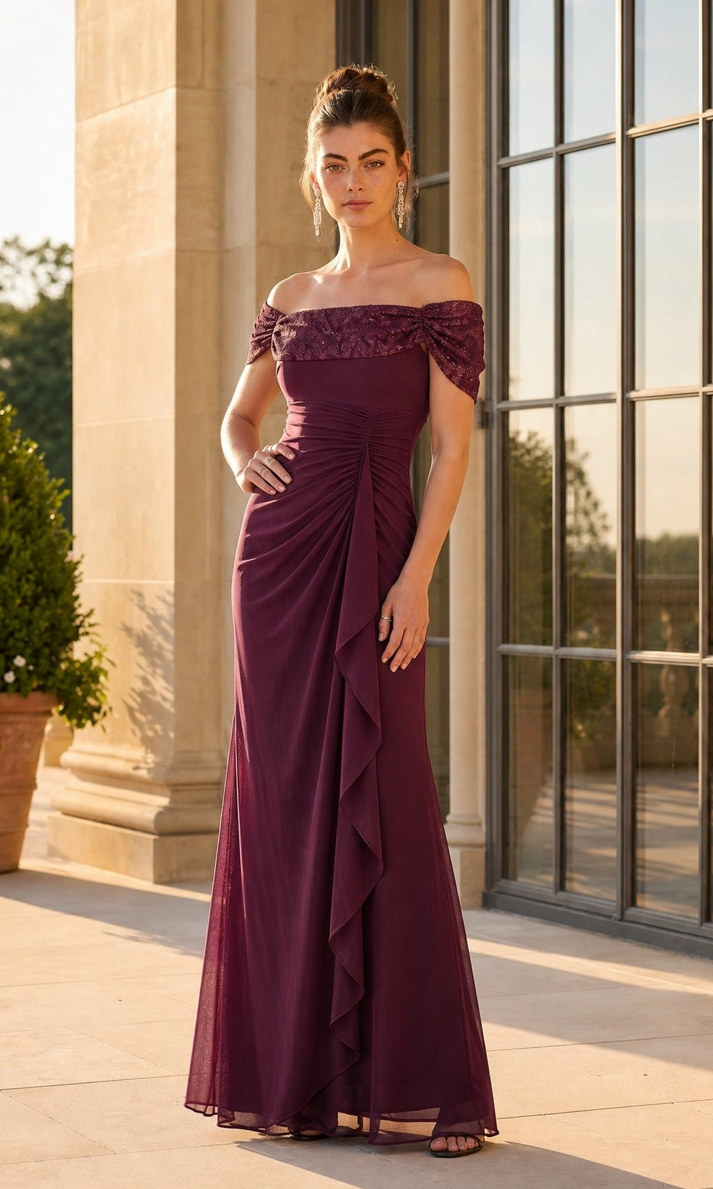 A woman stands outside wearing the Betsy & Adam A27436 long formal dress in wine, featuring an off-the-shoulder neckline, ruched details, and a ruffled front panel. Large windows and stone columns create a striking backdrop.