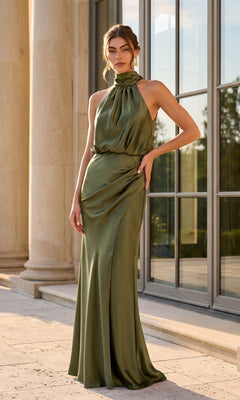Standing outside in front of tall windows, a woman wears the Betsy & Adam A28378 Long Formal Dress in olive. The floor-length sheath features a satin finish, halter neckline, and elegant draped details.