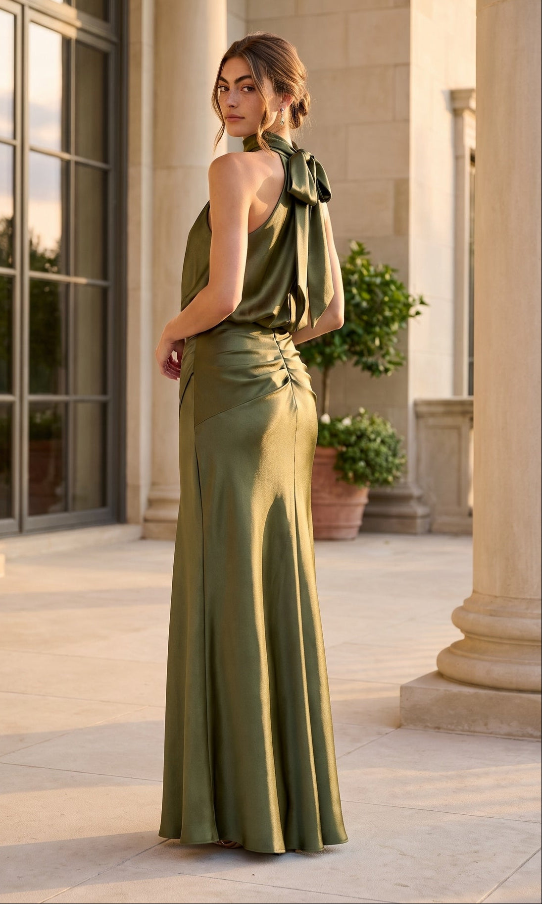 A woman wearing the Long Formal Dress: Betsy & Adam A28378—a flowing olive green satin gown featuring a bow on one shoulder—stands on a sunlit stone terrace near a column and potted plant.