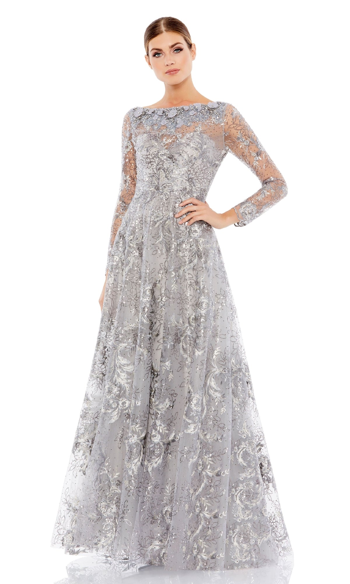 A woman poses with one hand on her hip in the Long Sleeve Long Formal Dress: Mac Duggal 67498, a floor-length silver evening gown with intricate floral lace patterns, set against a plain white background.