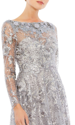A woman models the Mac Duggal 67498, a long sleeve formal dress with sheer fabric, silver embroidery, floral accents, and sequins, photographed from shoulders to knees against a white background.