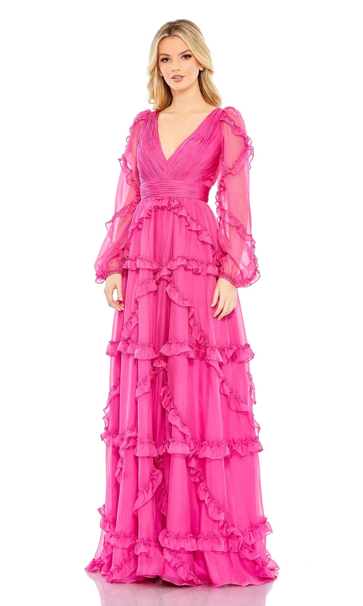 A woman stands against a white background wearing the Mac Duggal A68223 long sleeve formal dress—a floor-length, bright pink evening gown with ruffles and sheer sleeves.