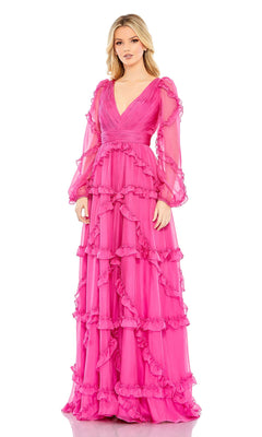 A woman stands against a white background wearing the Mac Duggal A68223 long sleeve formal dress—a floor-length, bright pink evening gown with ruffles and sheer sleeves.