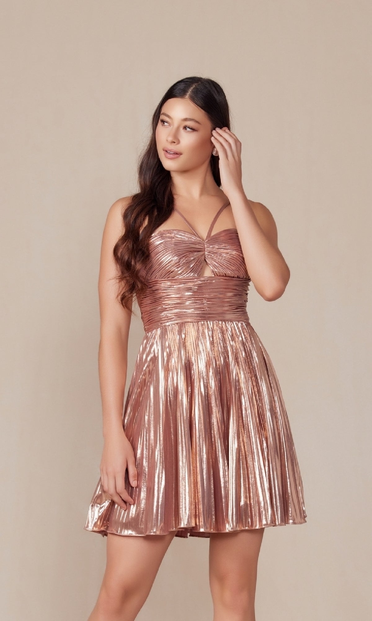 A woman stands against a plain background wearing the Nox Anabel A825 Pleated Metallic Homecoming Dress, featuring a sleeveless design, crisscross bodice detail, and a pleated skirt.