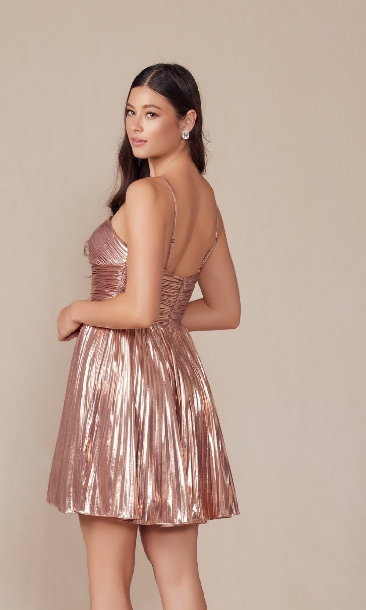 A woman stands against a plain light background, looking over her shoulder in the Pleated Metallic Homecoming Dress: Nox Anabel A825 in rose gold.