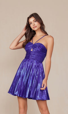 A woman wearing the Nox Anabel A825 Pleated Metallic Homecoming Dress, a shiny purple mini dress with spaghetti straps, stands against a plain beige background.