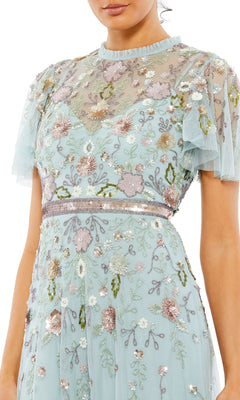 A woman wears the Mac Duggal Long Formal Dress 9136, featuring a light blue beaded A-line silhouette, multicolored floral embroidery, short sheer sleeves, and a high neckline.