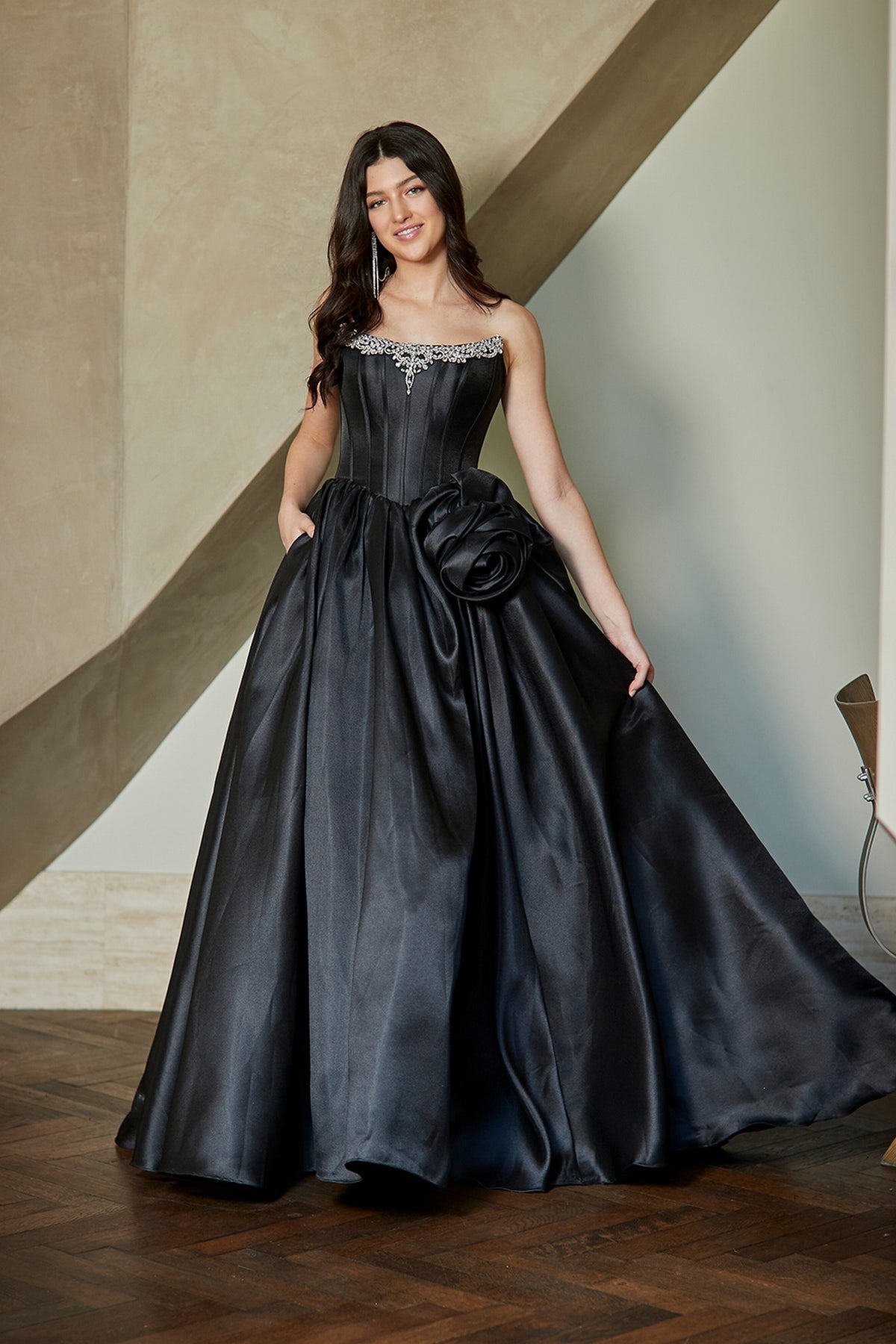 A person wearing a black strapless Corset Prom Ball Gown: Amelia Couture AL1401 with a rhinestone-embellished corset bodice and a large fabric flower accent on the skirt.