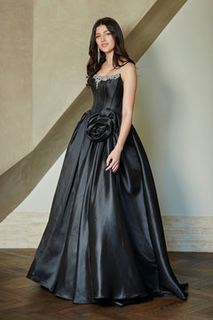 A person wearing the Amelia Couture AL1401 corset prom ball gown, a black strapless dress featuring a rhinestone-embellished neckline and a large floral accent at the waist.