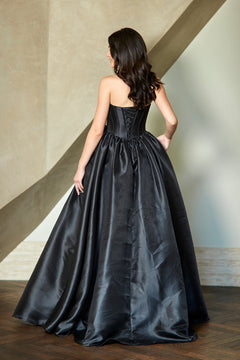 A woman wears the Corset Prom Ball Gown: Amelia Couture AL1401, a strapless black dress with a lace-up back and a full, floor-length skirt.