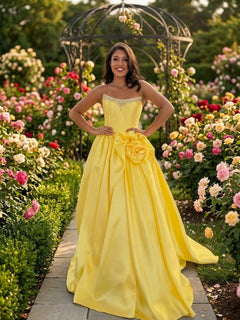 A woman wearing a yellow Corset Prom Ball Gown: Amelia Couture AL1401 with a strapless corset bodice and a large fabric flower on the waist, standing on a garden path lined with rose bushes.
