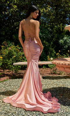A woman wears the Ruched Long V-Neck Prom Dress with Train 7107H in shiny light pink, featuring an open back and long train, as she stands on gravel near a stone fountain surrounded by lush greenery.