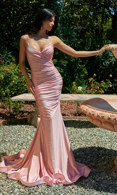 Wearing the Ruched Long V-Neck Prom Dress with Train 7107H, a woman stands outdoors on gravel near a stone fountain, surrounded by greenery. Her curve-hugging, floor-length pink gown beautifully accentuates her figure.