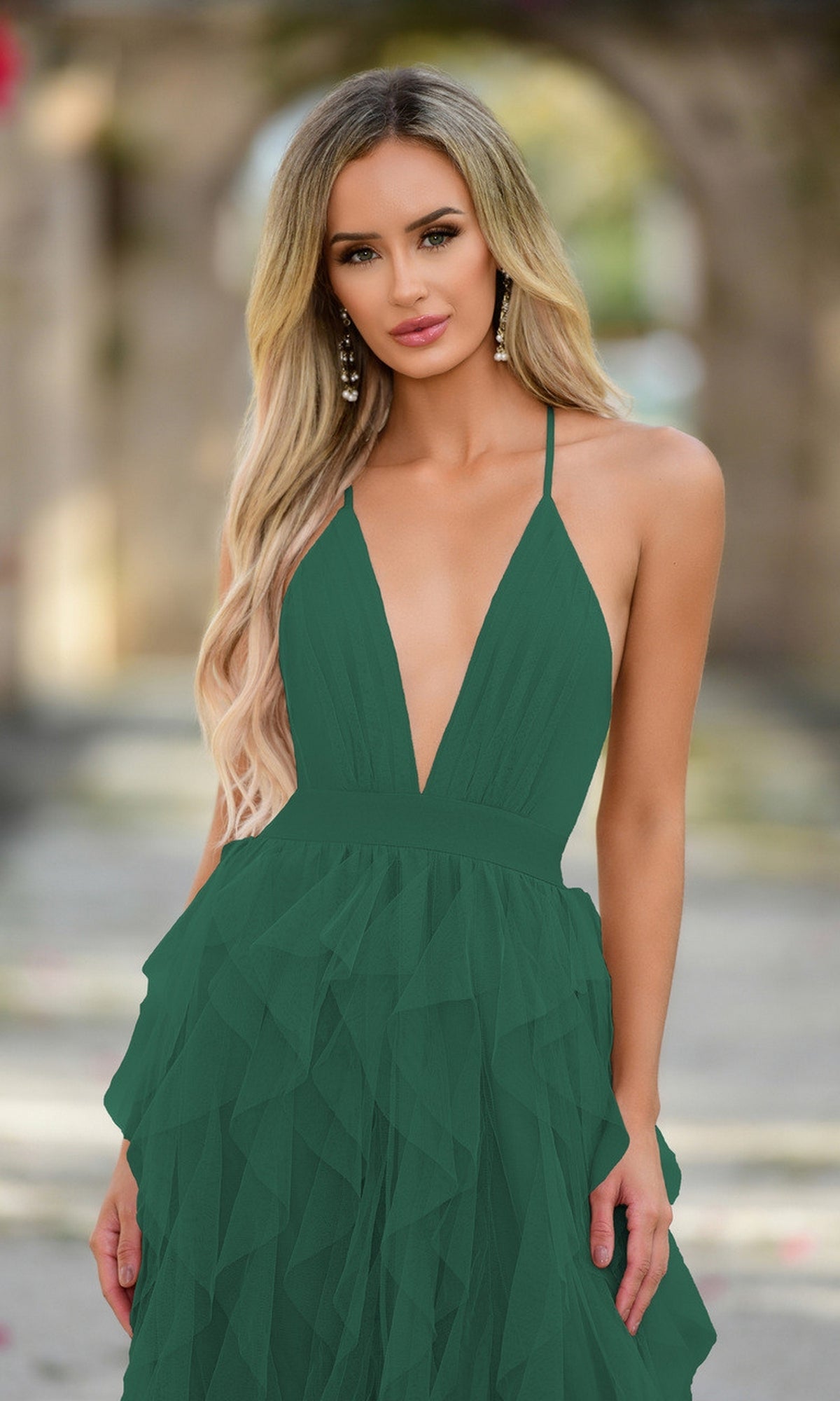 A woman with long blonde hair wears the Velvi Ruffled Long A-Line Prom Dress Alances in deep green, sleeveless style, standing outdoors with a blurred stone archway in the background.