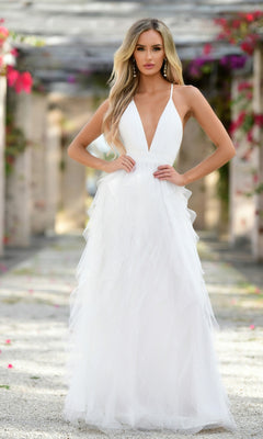 A woman stands outdoors among stone columns and greenery, wearing the Velvi Ruffled Long A-Line Prom Dress Alances, a sleeveless white gown with a deep V-neckline and layered tulle skirt.