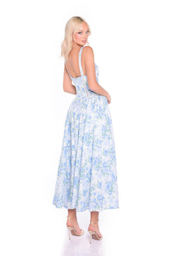A woman models the Anselma Blue Floral Cotton Summer Midi Dress with a fitted bodice and flared skirt, styled with nude heels against a white backdrop—ideal for wearing as a summer wedding dress.