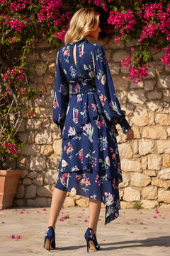 A woman in the Long-Sleeve Blue Floral Casual Midi Dress: Emery, featuring a smocked waistline and matching heels, stands outdoors on stone pavement, facing a stone wall topped with vibrant pink flowers.