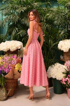 A woman in the Casual Midi Pink Floral Dress: Aria and beige heels stands among baskets of colorful flowers and greenery, glancing over her shoulder.