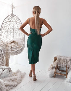 A woman with blond hair in a ponytail wears the Aspen Sleek Midi Slip Party Dress and heels, standing in a white room with woven furniture and textured blankets.