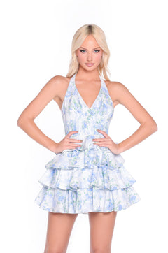A woman stands with hands on hips, wearing the Aurelia Mini Blue Floral Halter Summer Dress featuring a sleeveless design, blue and white floral pattern, and a tiered mini skirt.