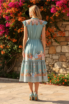 A woman stands outdoors with her back to the camera, in front of flowers and a stone wall, wearing the Ava Dusty Teal Floral Casual Midi Dress with a belted waist and matching shoes.