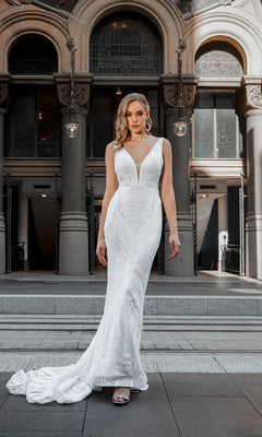 A woman stands outdoors before arched columns, wearing the Tina Holly BA109 white lace mermaid wedding gown—a fitted, sleeveless dress with a long train.
