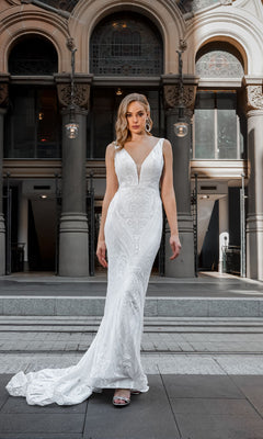 A woman wears the White Lace Mermaid Wedding Gown: Tina Holly BA109, featuring a long train, as she stands on stone steps before an ornate building.