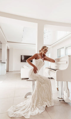 A woman in the Tina Holly BA999 White Lace Bodycon Bridal Gown sits on a white stool next to a white grand piano in a bright, modern living room.