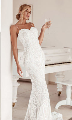A woman in the White Lace Bodycon Bridal Gown: Tina Holly BA999 stands next to a white piano, holding a glass of wine and gazing away from the camera.
