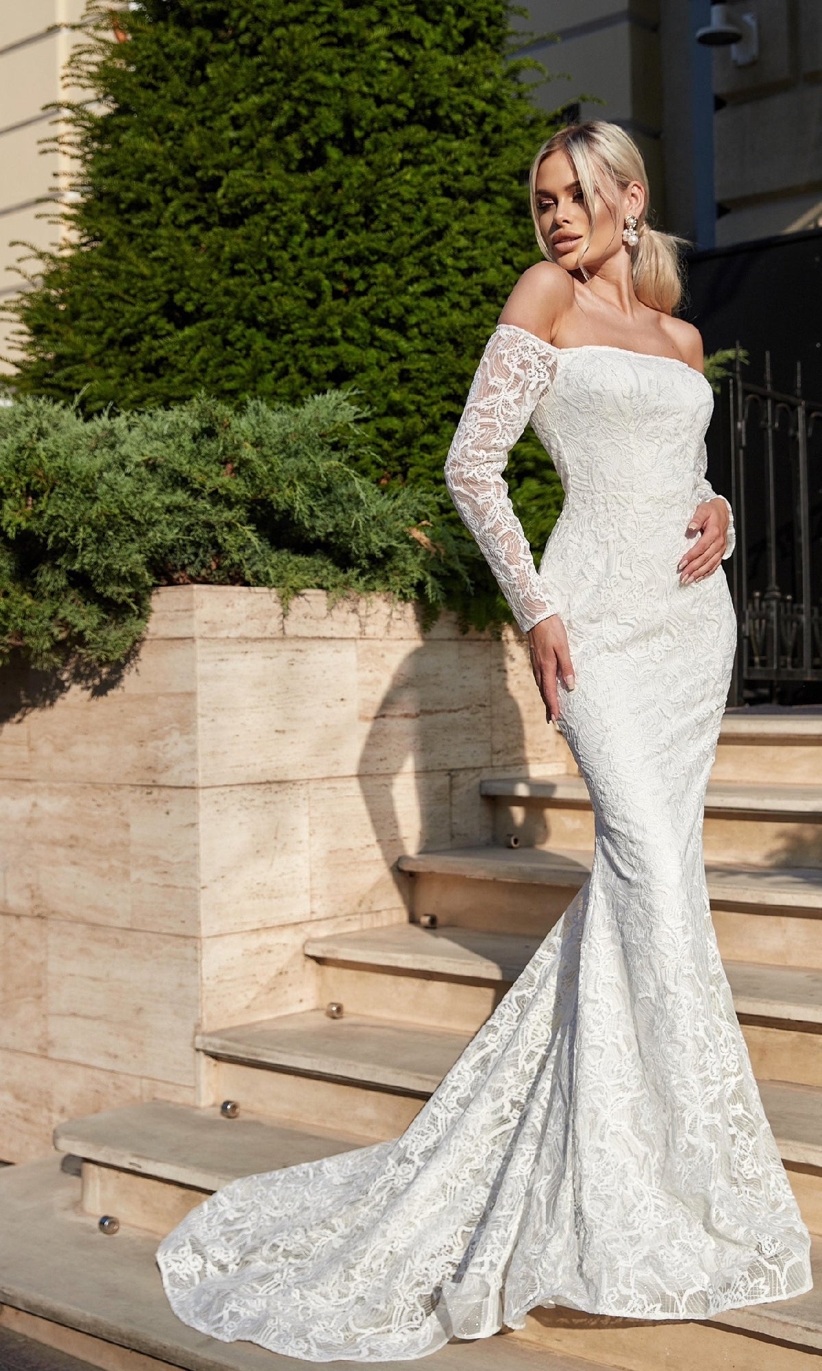 Wearing the Tina Holly BB007 Long Lace White Formal Gown, a woman stands gracefully on outdoor stone steps beside lush green shrubs.