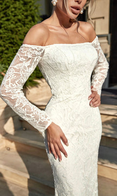 A woman stands outdoors near steps, surrounded by lush greenery, wearing the Long Lace White Formal Gown: Tina Holly BB007, featuring intricate white lace and an off-the-shoulder design.