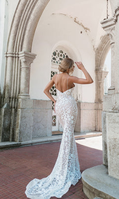 A woman in the Tina Holly BB008 long white lace formal dress stands outdoors under a stone arch, facing away and adjusting her hair.