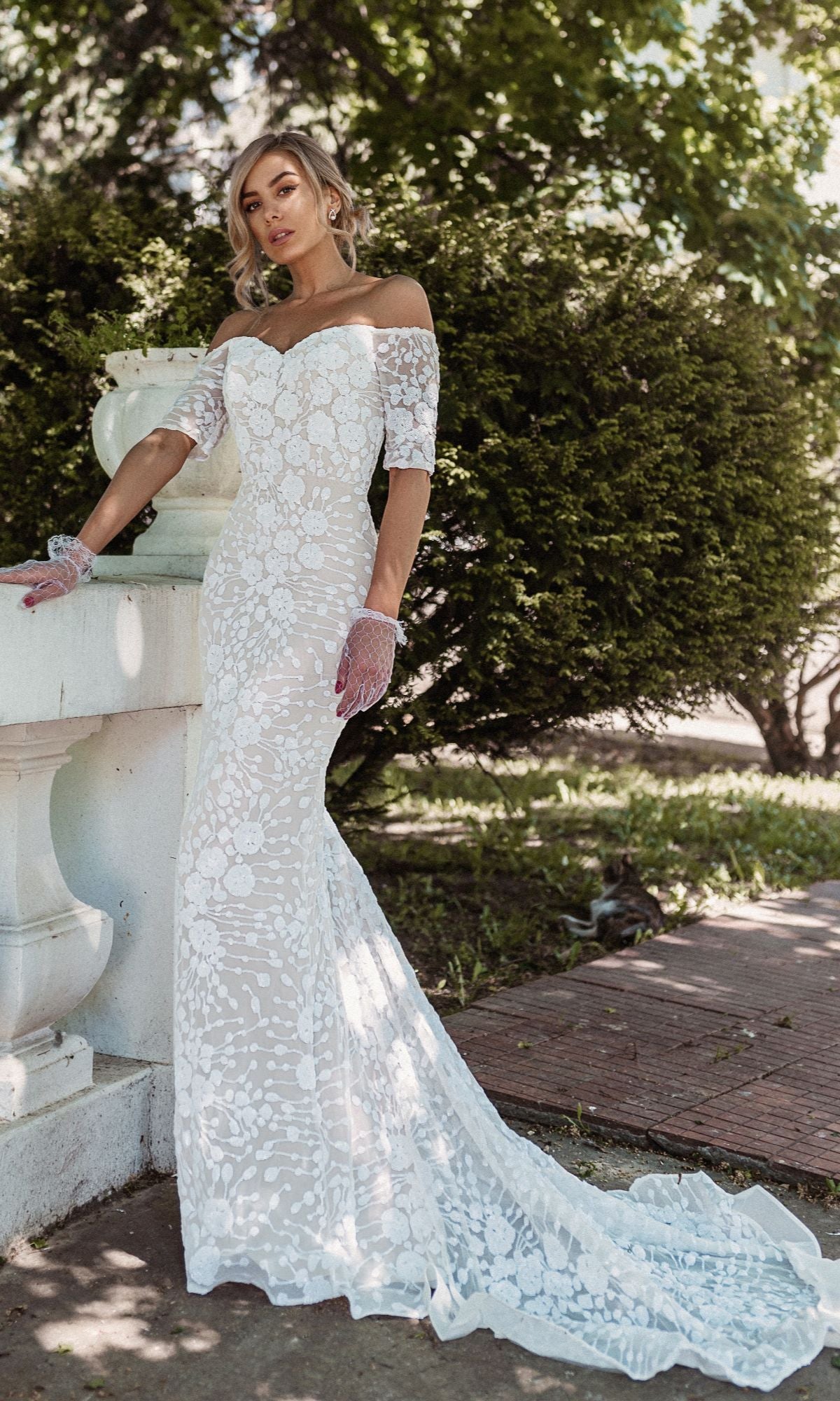 A woman wearing the Off-Shoulder White Bridal Gown: Tina Holly BB009, featuring intricate lace and a long train, stands outdoors near a stone railing and lush greenery.