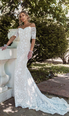 A woman wearing the Off-Shoulder White Bridal Gown: Tina Holly BB009, featuring intricate lace and a long train, stands outdoors near a stone railing and lush greenery.