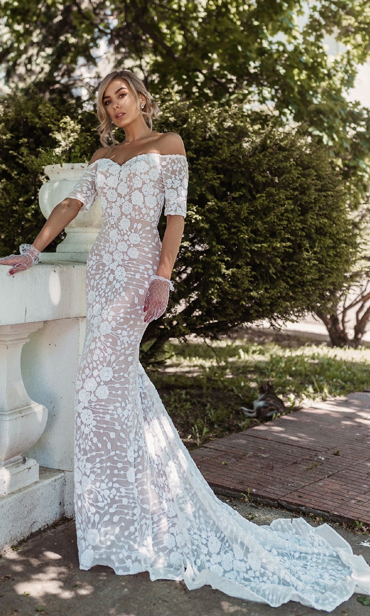 A woman stands outdoors by lush greenery, wearing the Off-Shoulder White Bridal Gown: Tina Holly BB009, which features a long train and sheer floral-patterned details.