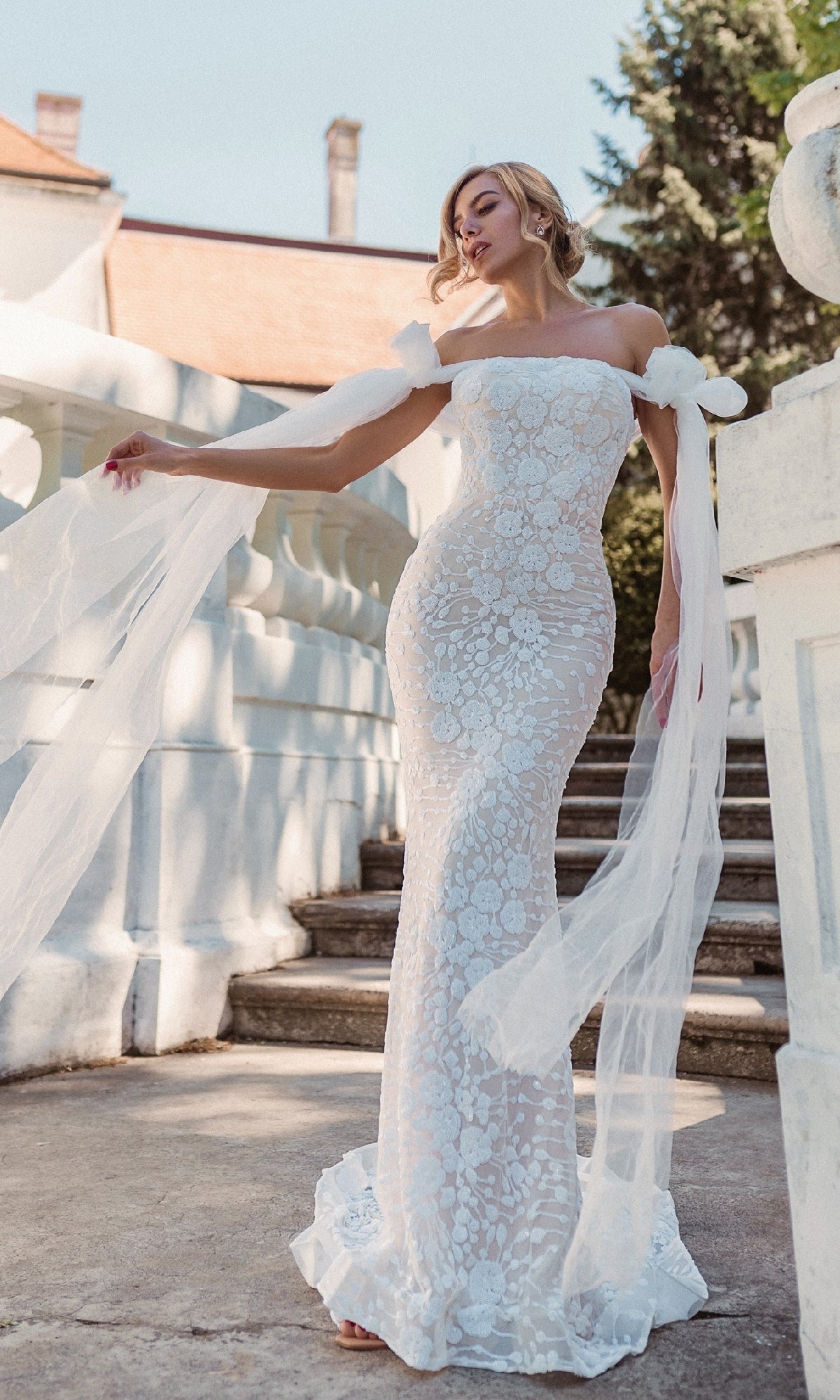 A woman poses outdoors on stone steps beside a white balustrade, wearing the Tina Holly BB010 Strapless White Wedding Gown.