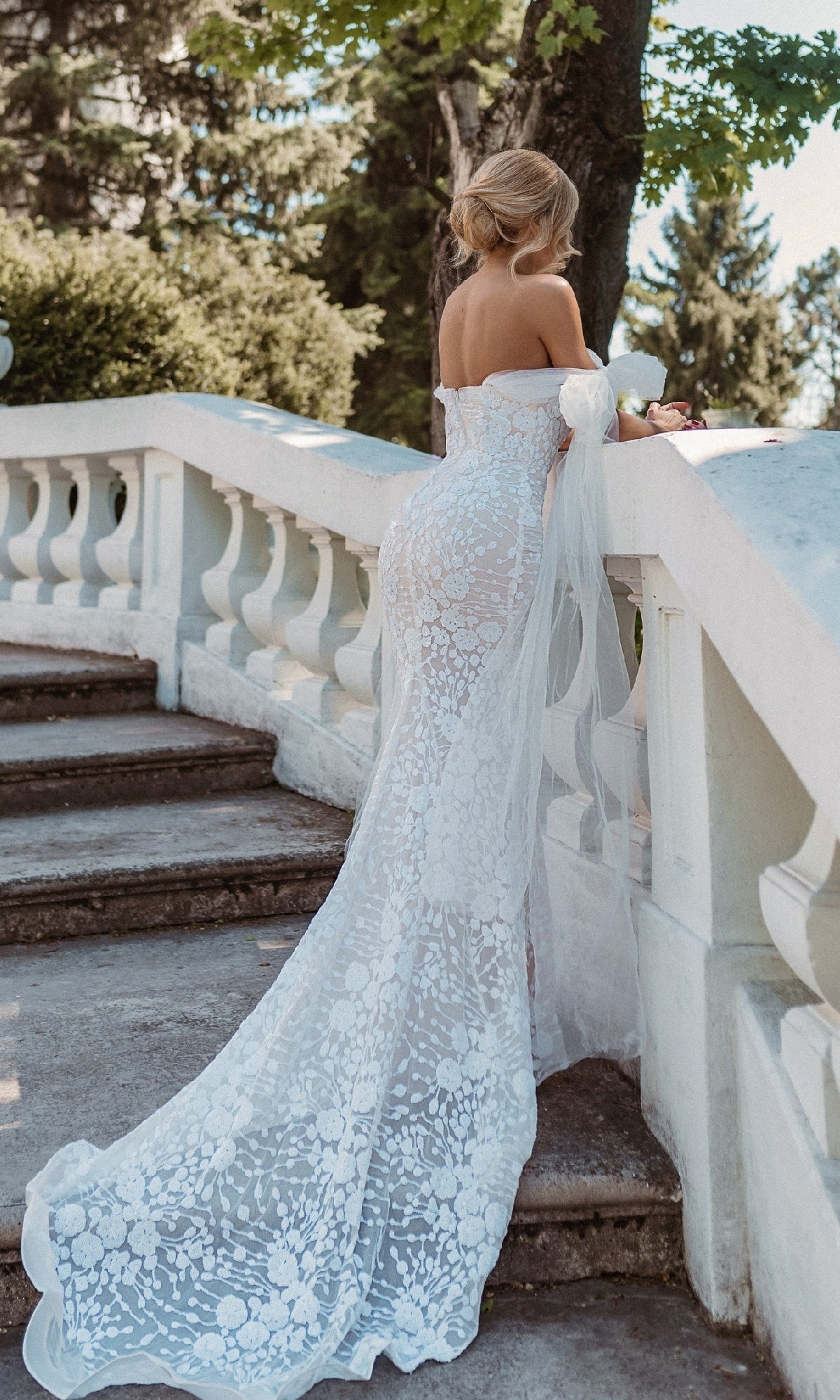 A woman in the Strapless White Wedding Gown: Tina Holly BB010 stands on stone steps outdoors, facing away and leaning on a white balustrade.