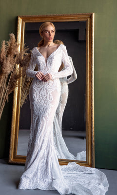 A woman in the Tina Holly BB021 long sleeve white bridal gown stands before a large gold-framed mirror, her elegant dress and its reflection beautifully displayed behind her.