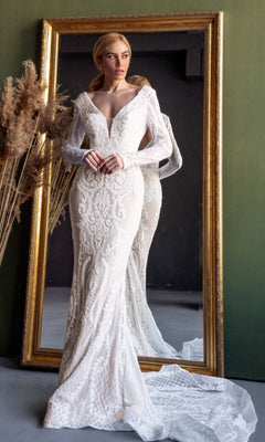 A woman wearing the Tina Holly BB021 long sleeve white bridal gown with a mermaid skirt stands before a large ornate mirror, dried plants on the side.