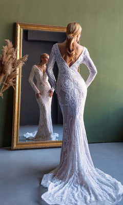 A woman wearing the Long Sleeve White Bridal Gown: Tina Holly BB021 stands facing a mirror, her reflection visible, in a room with a green wall and dried plants.