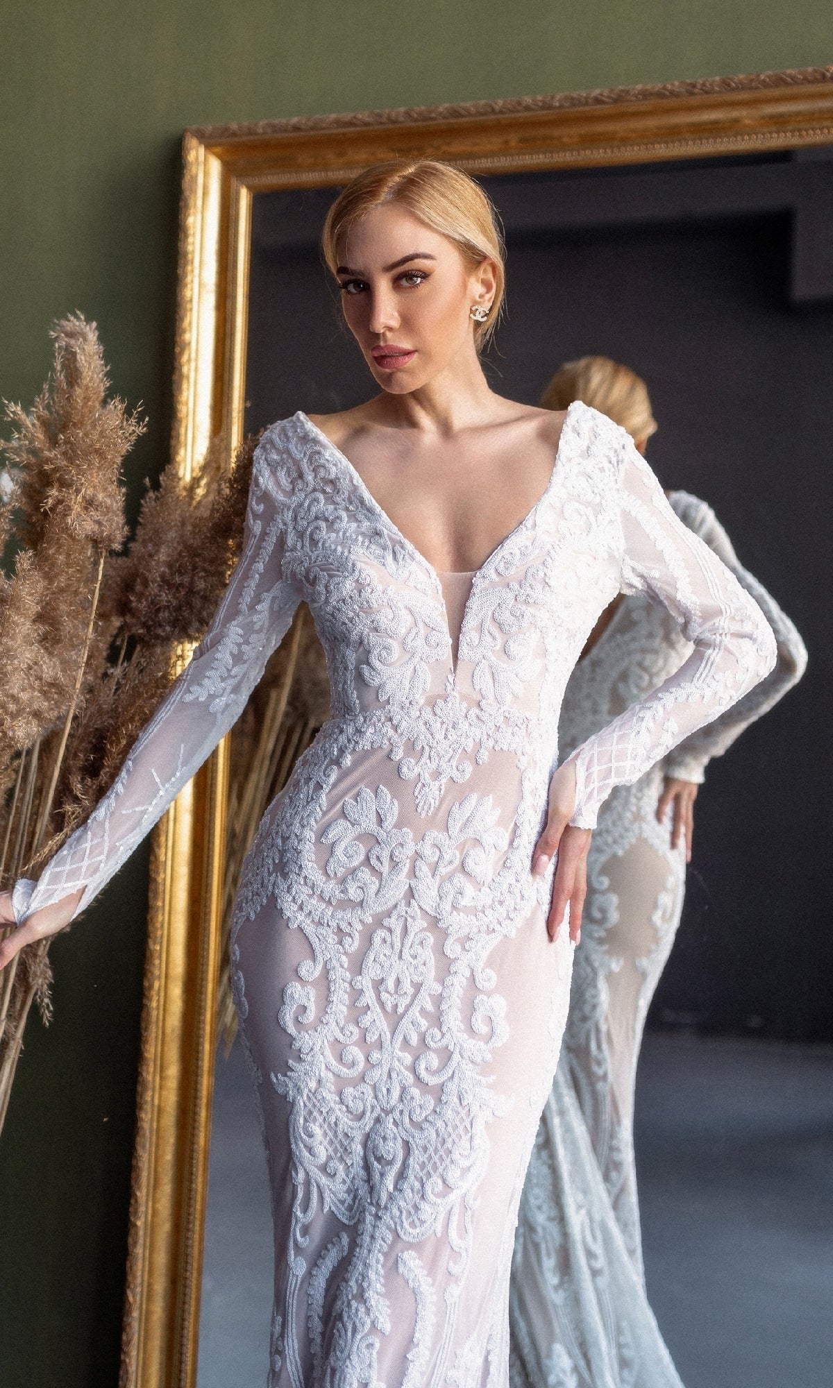 A woman wearing the Long Sleeve White Bridal Gown: Tina Holly BB021 stands before a large mirror with dried pampas grass at her side.