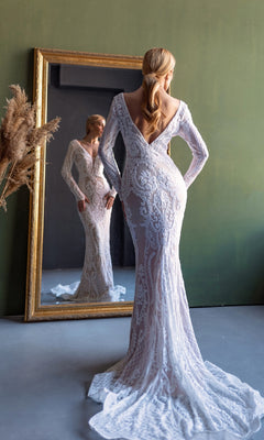 A woman with long hair in a low ponytail wears the Tina Holly BB021 long sleeve white bridal gown, featuring fitted lace and a deep V-back, as she stands before a large mirror.