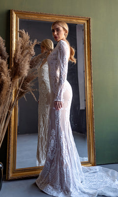 A woman wearing the Long Sleeve White Bridal Gown: Tina Holly BB021 stands before a large framed mirror, with tall dried grasses in a vase nearby.