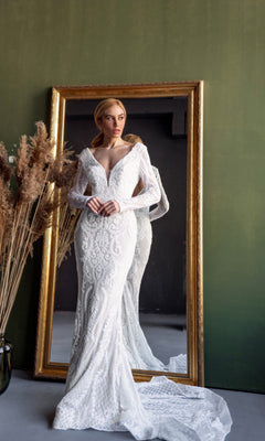 A woman in the Long Sleeve White Bridal Gown: Tina Holly BB021 stands before a large gold-framed mirror set against a green wall, with dried pampas grass arranged in a vase nearby.
