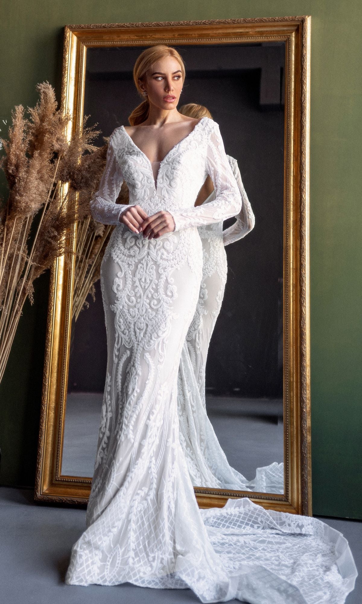 A woman wearing the Tina Holly BB021 Long Sleeve White Bridal Gown stands before a large ornate mirror, with dried pampas grass nearby.