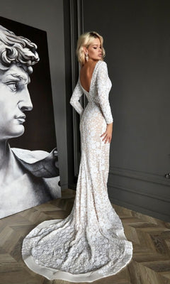 A woman in the Tina Holly BB023 Long Formal Dress, featuring long sleeves and a backless lace overlay, stands indoors beside black-and-white classical statue artwork.