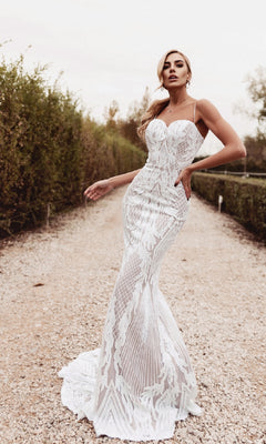 A woman stands on a gravel path outdoors, posing with one hand on her hip in the Long White Mermaid Wedding Dress: Tina Holly BB050—a strapless, form-fitting lace bridal gown with intricate patterns.