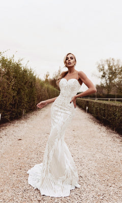A woman stands outdoors on a gravel path wearing the Tina Holly BB050, a long white mermaid wedding dress featuring intricate patterns, a flared hem, and a form-fitting sweetheart neckline.